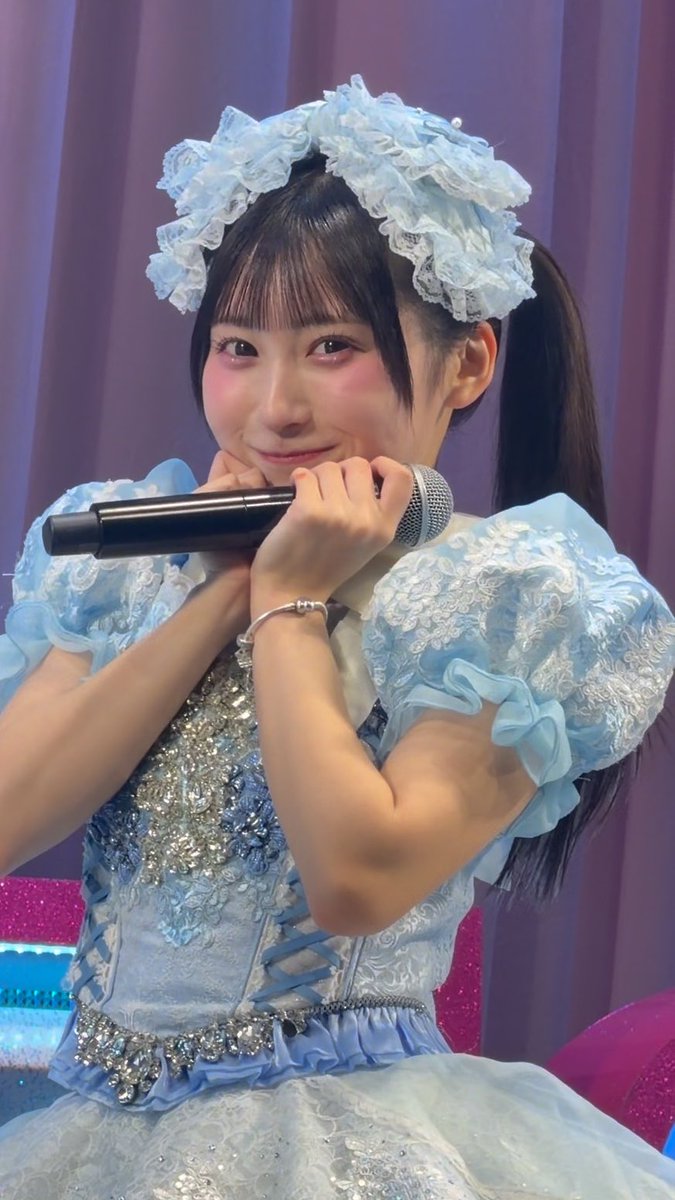 A close-up of a Japanese pop idol wearing an elaborate light blue frilly costume with sequin details and a matching blue lace headpiece, holding a microphone while smiling at the camera during what appears to be a fan event.
