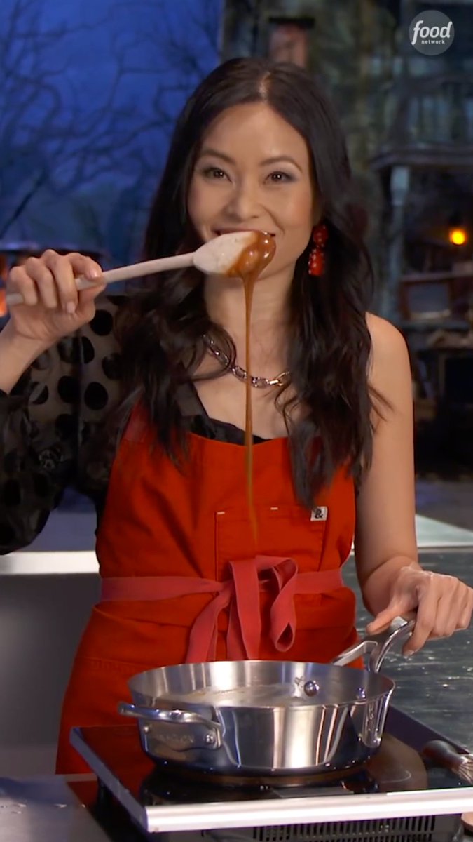 A woman wearing a red apron with an ampersand ("&") symbol smiles while lifting a wooden spoon from a stainless steel pot, letting caramel drip from it. The Food Network logo appears in the top right corner, tying into the post's promotion of Halloween Wars, where chef Shinmin demonstrates homemade caramel.