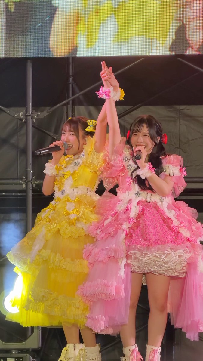 Two performers in elaborate, ruffled yellow and pink dresses sing into microphones and form a heart shape with their arms on stage.