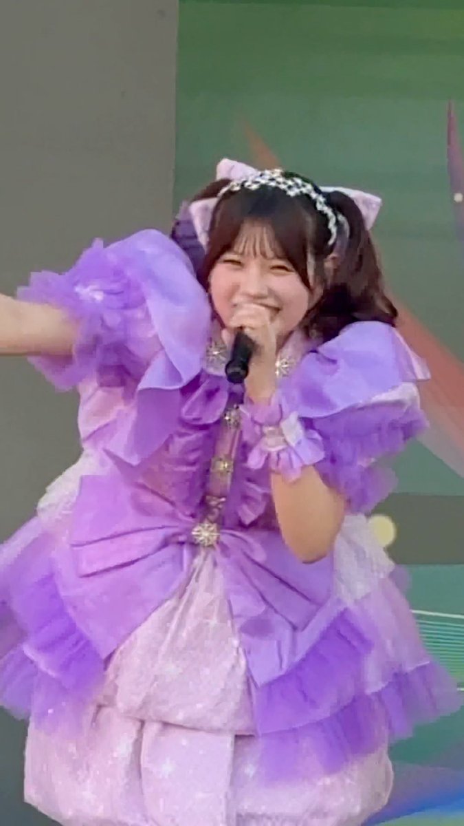 A performer singing into a microphone while wearing a frilly purple and white dress, smiling brightly at the camera.