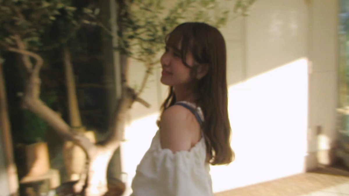 A woman with long brown hair wearing a white off-shoulder top smiles softly while standing outdoors with sunlight filtering through nearby foliage.