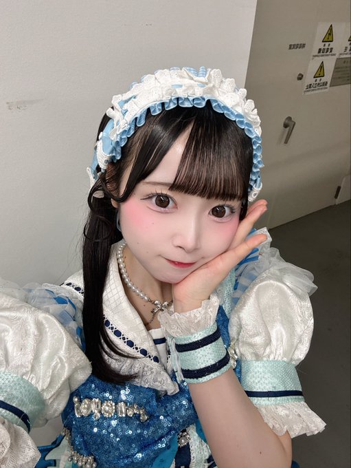 A selfie of a young woman in a blue and white frilly maid-style outfit with a matching headband and pearl necklace, resting her face on her hand.