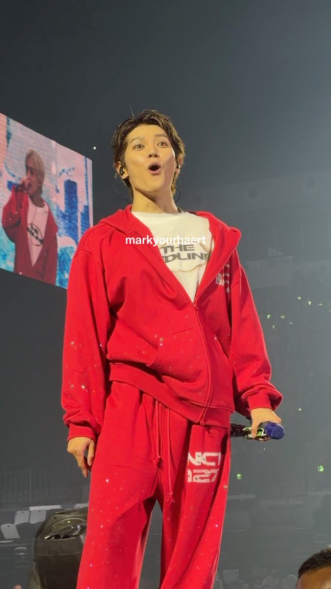 A K-pop performer in a red zip-up hoodie and matching pants performs on stage, holding a blue microphone with a surprised expression. A watermark reads "markyourhaert".