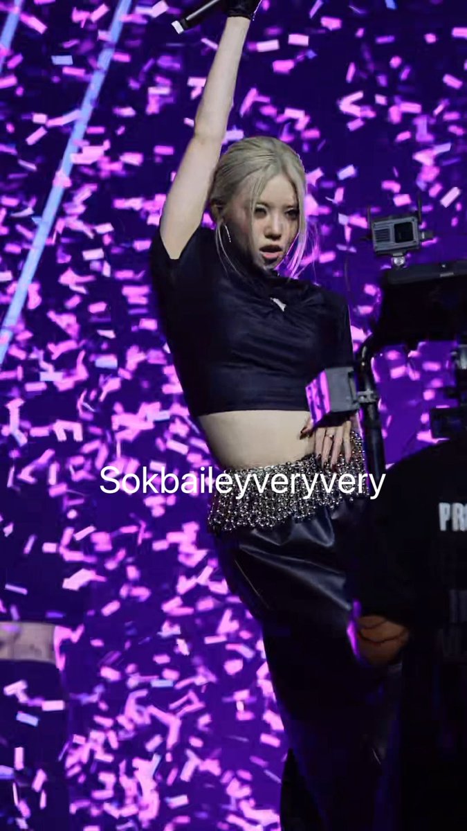 A blonde performer with her arm raised triumphantly stands on stage amid a shower of purple confetti, wearing a black crop top and embellished skirt. The watermark "Sokbaileyyvery" appears across the image, and the post celebrates Bailey from AllDay Project (올데이프로젝트), matching the caption's enthusiastic fandom pride.