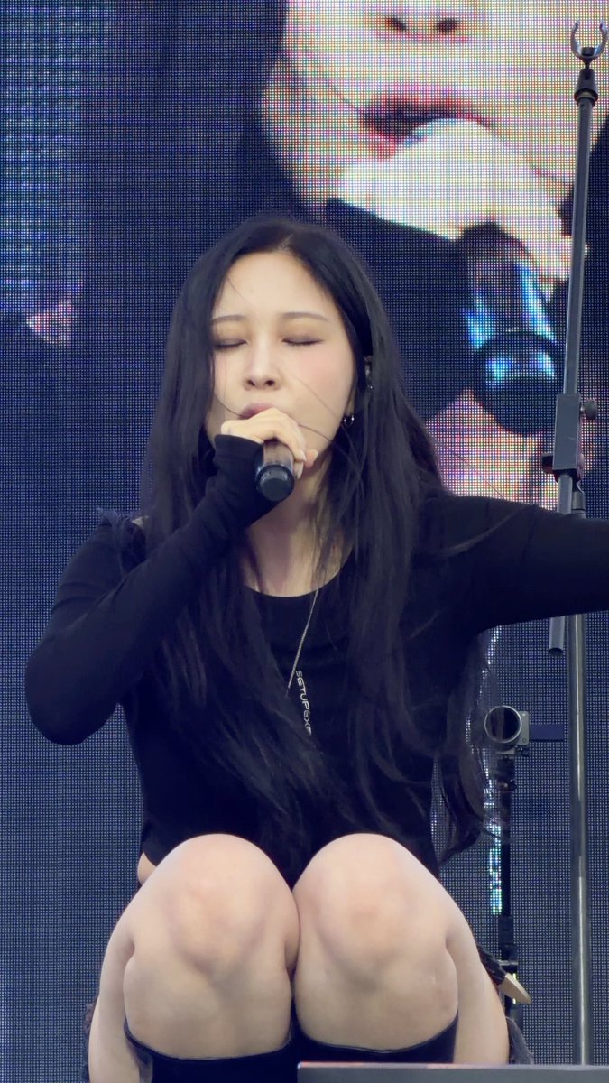 A female performer with long black hair wearing a black long-sleeved outfit and thigh-high boots, kneeling on stage while holding a microphone with her eyes closed.