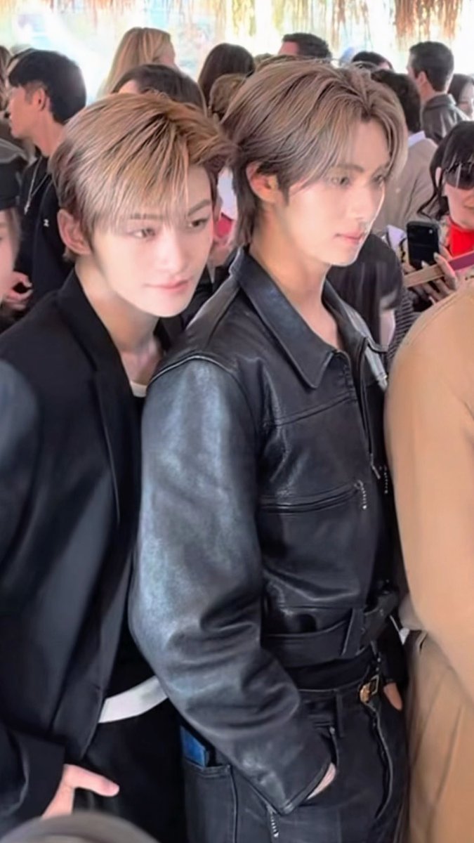 Two young men with blonde hair pose closely together in a crowded outdoor setting, both wearing dark outfits including a black leather jacket.