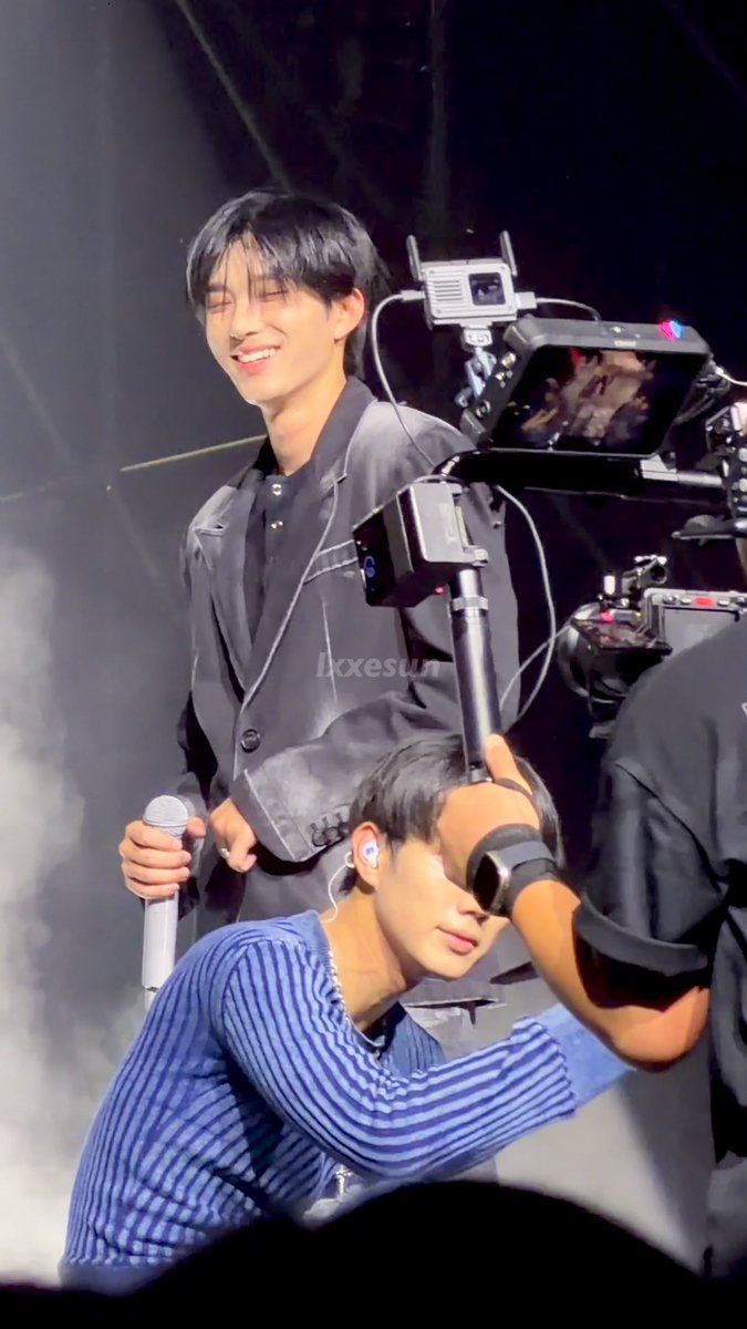 Two young men perform on stage during what appears to be a concert, with one holding a microphone and smiling while a professional camera is being operated nearby.