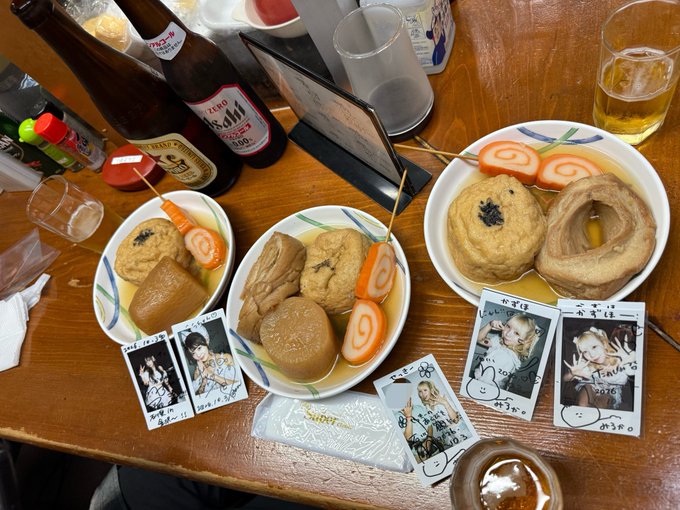 A Japanese meal of oden featuring fish cakes, daikon, and egg is served alongside beer bottles and polaroid photos of idols (including one labeled '2014.7.31 かな') signed by fans.