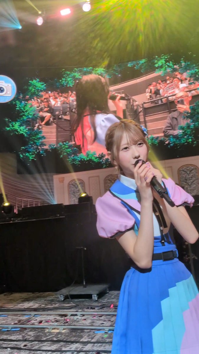 A young performer in a blue and pink outfit singing into a microphone on stage with colorful lighting and a large screen behind showing the audience.