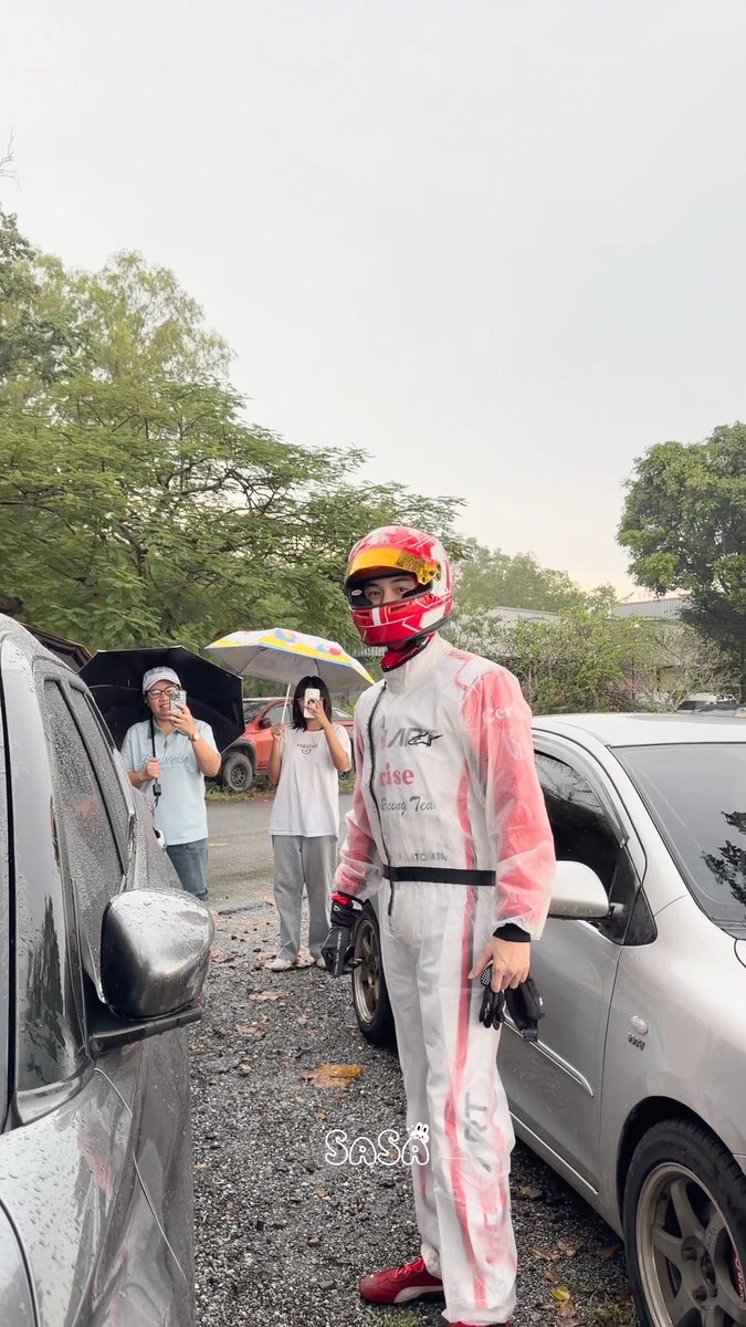 A person dressed in a white and red racing suit with a helmet, standing in the rain between two cars while two others watch.