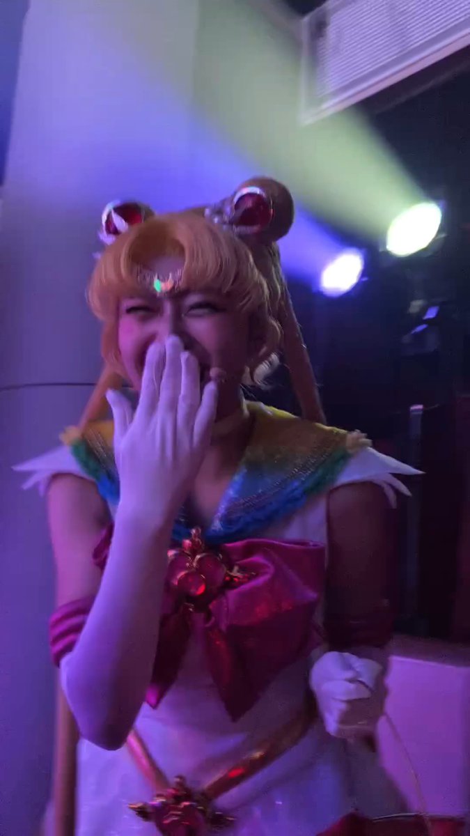 A cosplayer dressed as Sailor Moon (with blonde odango buns, a tiara, and a white sailor fuku with a red bow) laughs while covering her mouth with her gloved hand, bathed in colorful stage lighting. The caption expresses gratitude for receiving a second treasured item, and the joyful cosplayer's reaction captures that same sense of excitement and happiness.