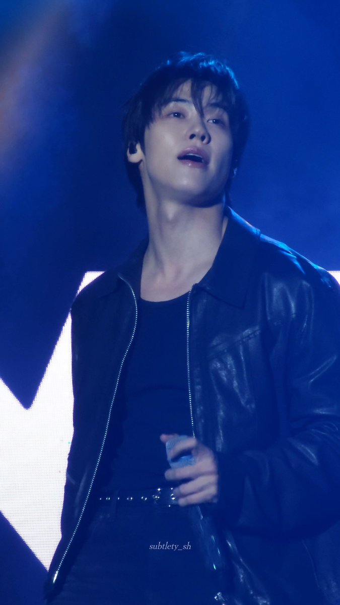 A young male performer on stage in a black leather jacket holding a microphone, with the watermark 'subtlety_sh' visible.