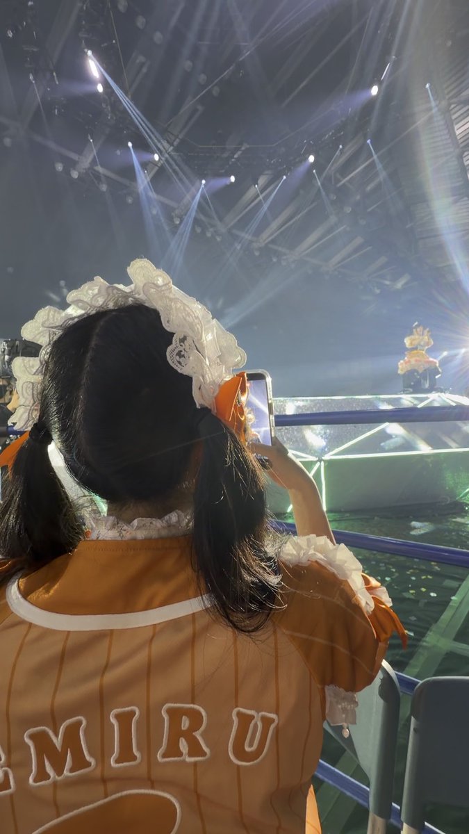 A person wearing an orange and white maid-style outfit with 'MIRU' printed on the back and a white lace headband, holding up a smartphone to film a stage performance with bright spotlights.