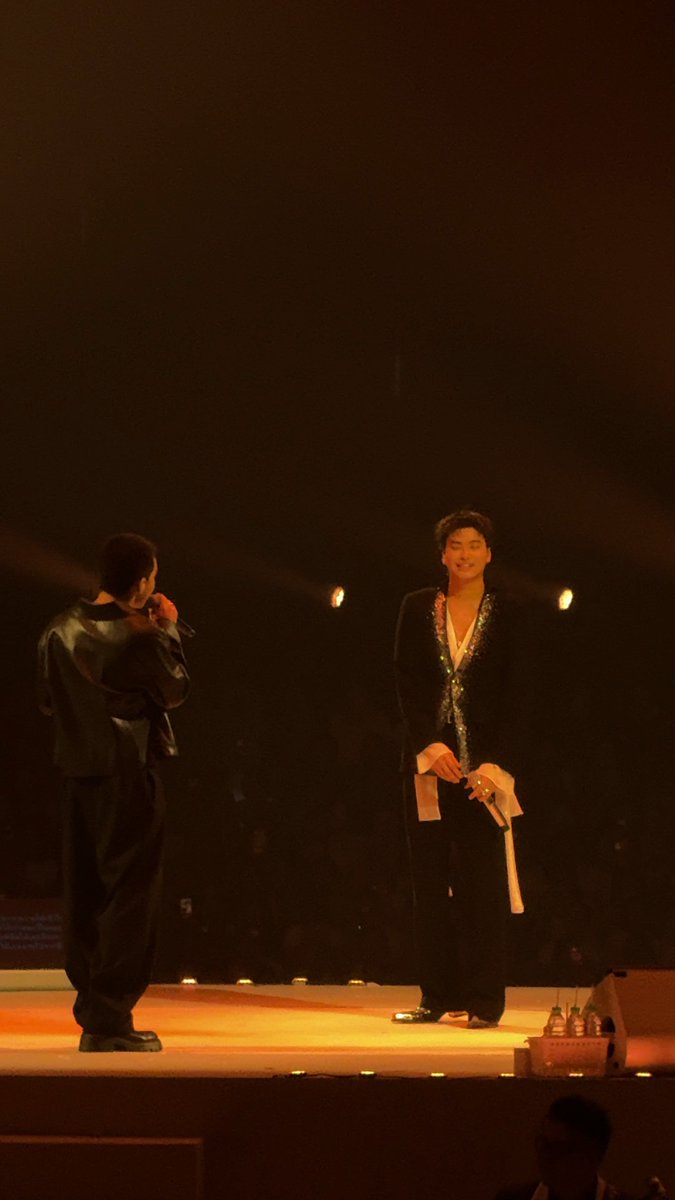 Two men stand on a dimly lit stage, with the man on the right wearing a sparkling green-trimmed jacket and holding a white towel.