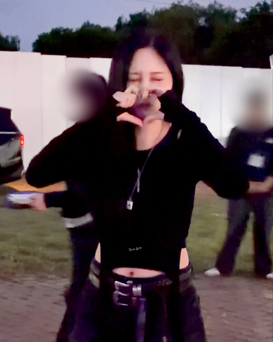 A young woman with dark hair stands outdoors at dusk, wearing a black top with an arm warmer, forming a heart shape with her hands over her face, while two blurred figures stand behind her on the grass.