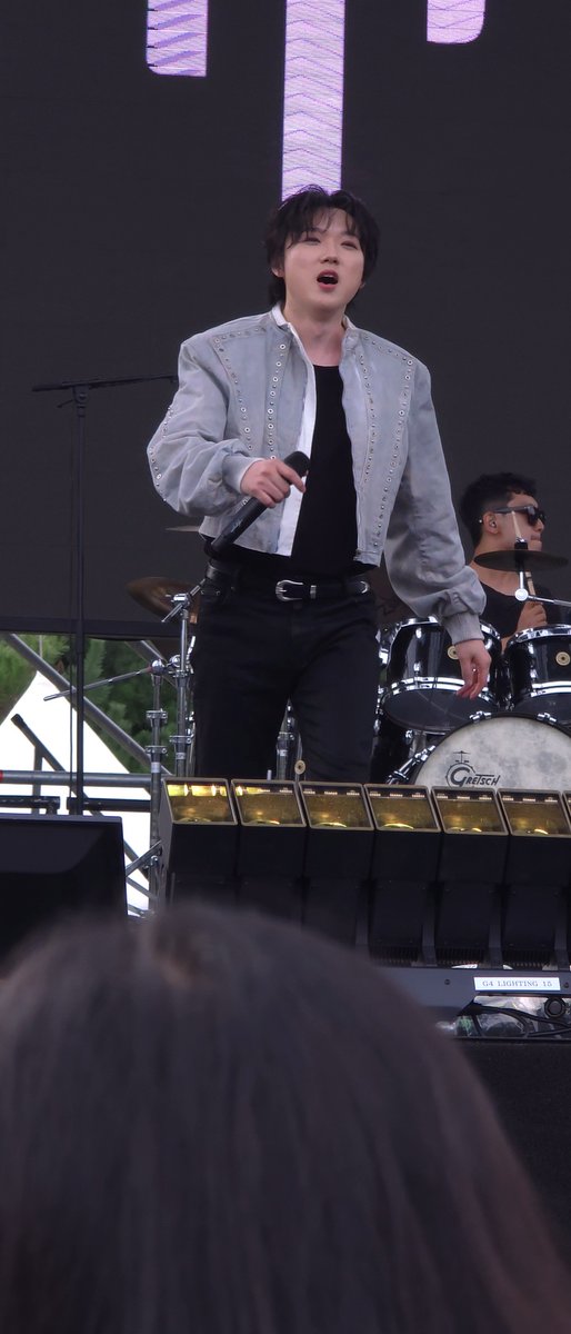 A young male performer sings into a microphone on stage, wearing a light gray embellished jacket over a black shirt. A drum set and audience members are visible in the background of this outdoor concert.