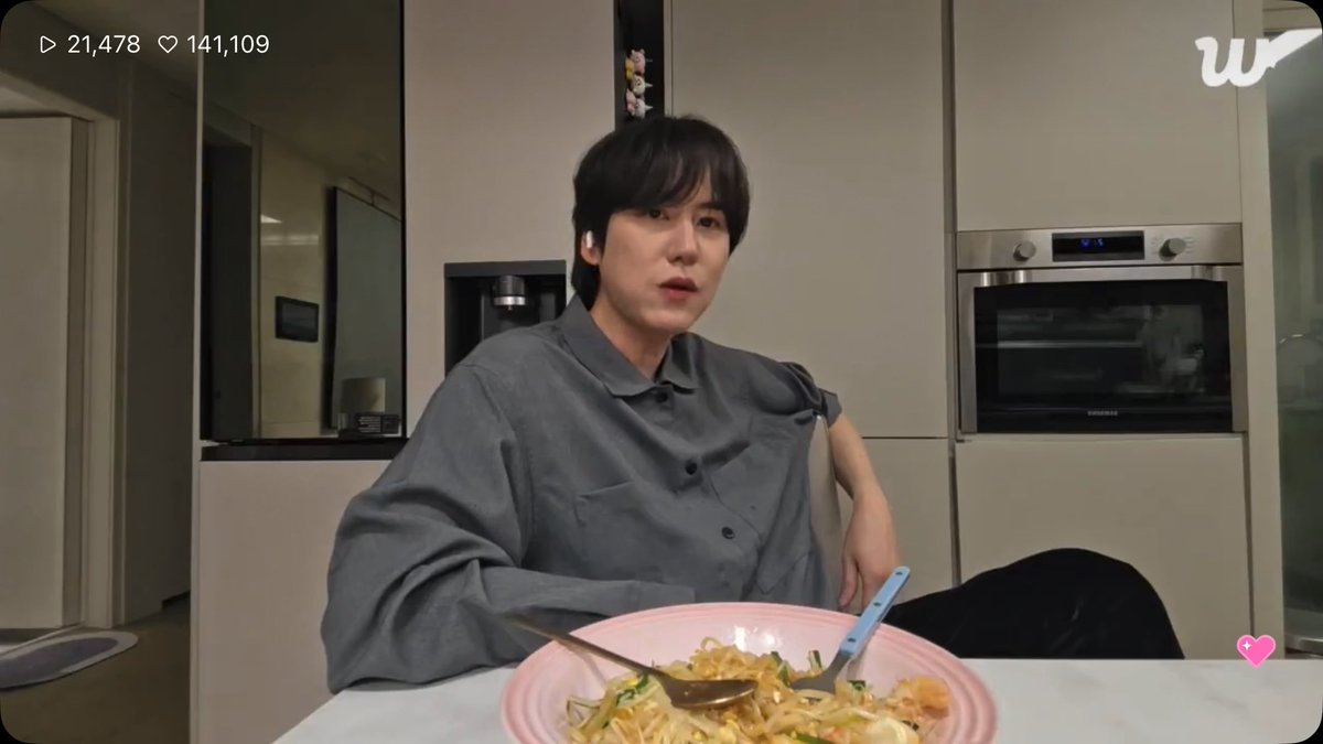 A screenshot from a Weverse livestream showing a young man with dark hair in a gray shirt sitting at a kitchen table with a pink bowl of noodles, with 21,478 views and 141,109 likes.