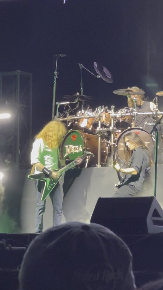 A live concert performance featuring guitarists on stage with a drum kit displaying the 'MEGADETH' logo in the background.