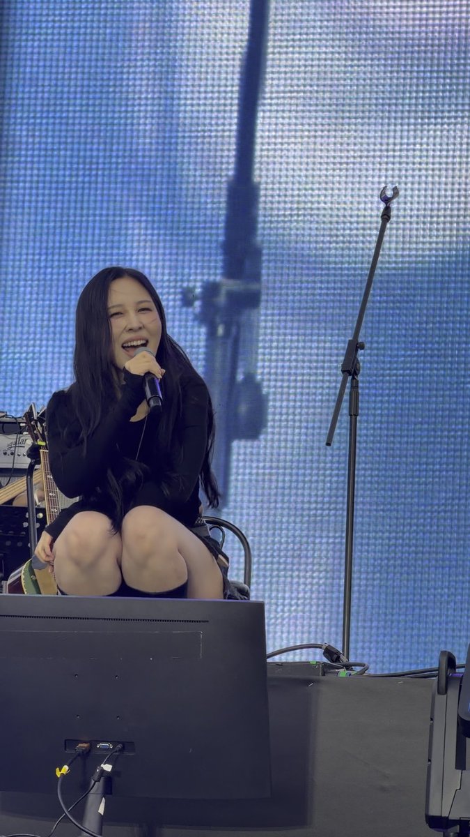 A woman with long dark hair, wearing a black outfit, sits on stage holding a microphone and smiling, with a large blue LED screen behind her.