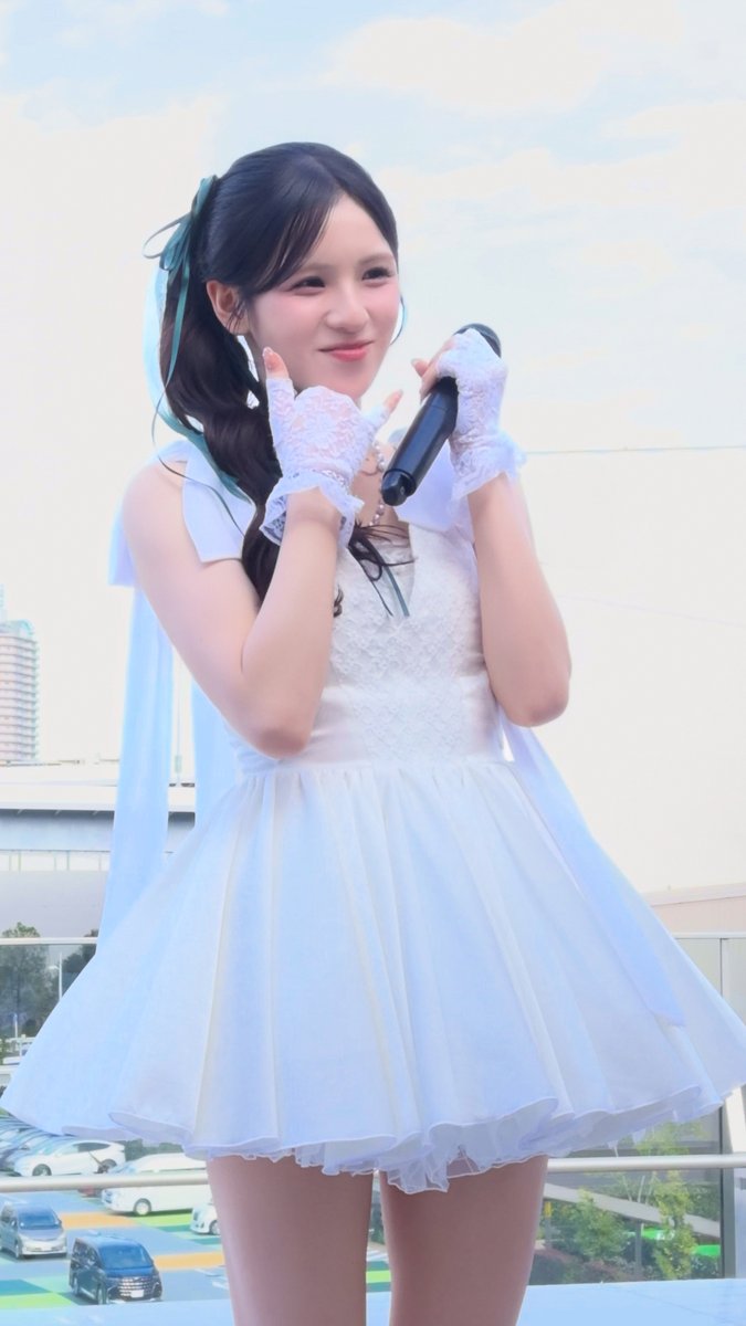 A young woman in a white frilly dress with lace gloves, holding a microphone and posing on an outdoor stage.