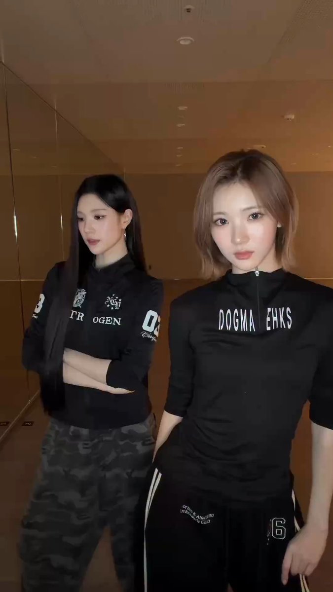 Two young women pose together in athletic wear; one wears a black shirt with 'STR OGEN' and camo pants, while the other wears a black top reading 'DOGMA EHKS' and black pants.