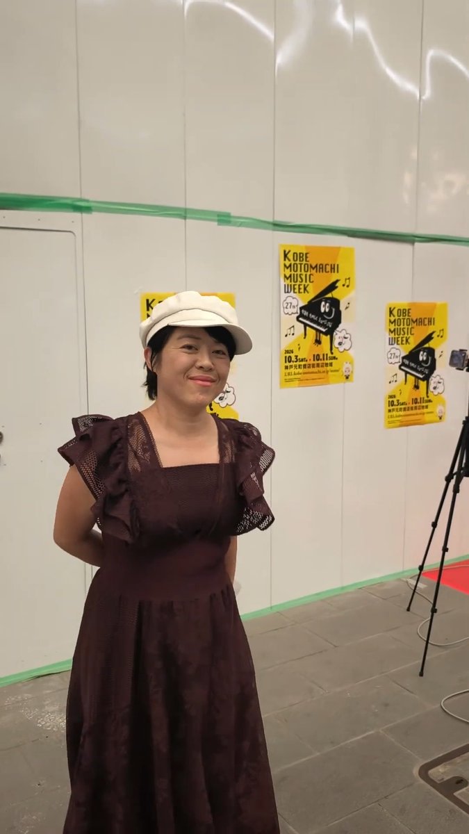 A woman wearing a white cap and dark brown ruffled dress poses in front of yellow posters for the "KOBE MOTOMACHI MUSIC WEEK" event held from October 3-5 and 10-11.