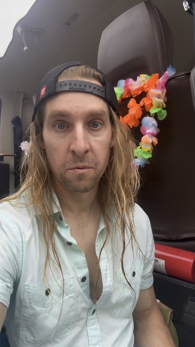 A selfie of a man with long blonde hair wearing a black cap and light blue shirt, sitting in what appears to be a vehicle with colorful flower leis hanging behind him.