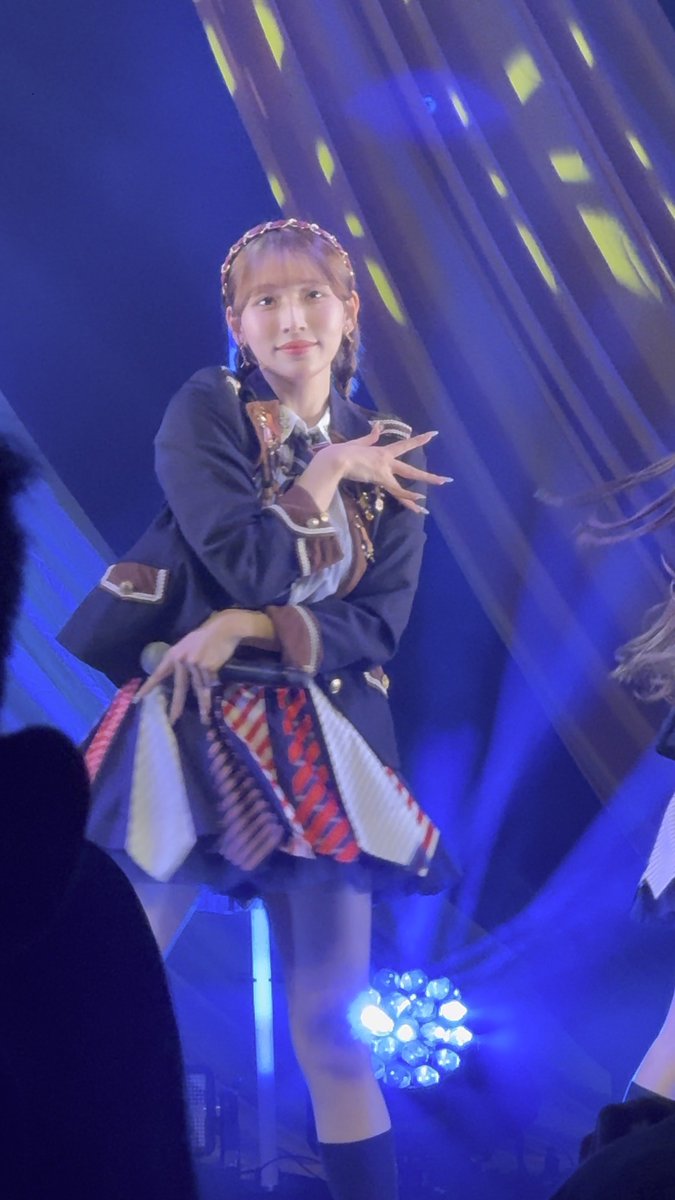 A young woman performs on stage under blue lighting, wearing a dark blazer and a pleated skirt.