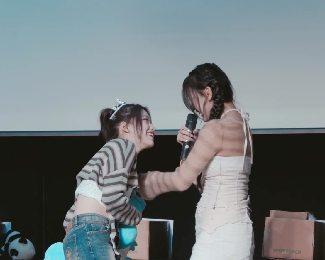 Two women share a playful, close moment on stage at what appears to be a fan meeting, with one holding a microphone while the other leans in with a bright smile. Pink gift bags labeled "KPOP MERCH" are visible in the foreground, fitting the context of a casual K-pop fan event interaction suggested by the caption's playful dialogue ("Kayoung-ah~ Huh? Ah~ no... Huh?").