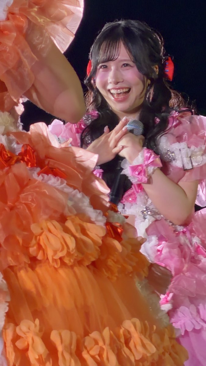 A smiling young woman with pigtails in a pink ruffled idol outfit, holding a microphone while standing behind a performer in an orange ruffled costume.