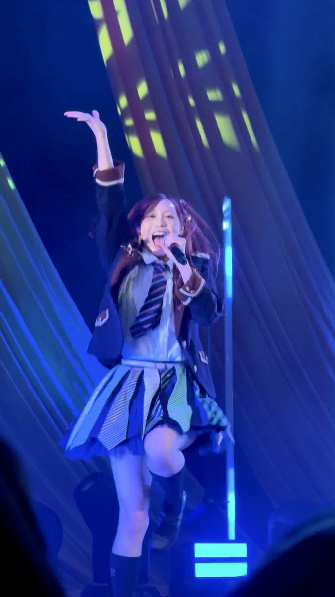 A young performer with pigtails sings into a microphone on a blue-lit stage, wearing a school uniform-style outfit with a striped tie, raising one arm in an energetic pose.
