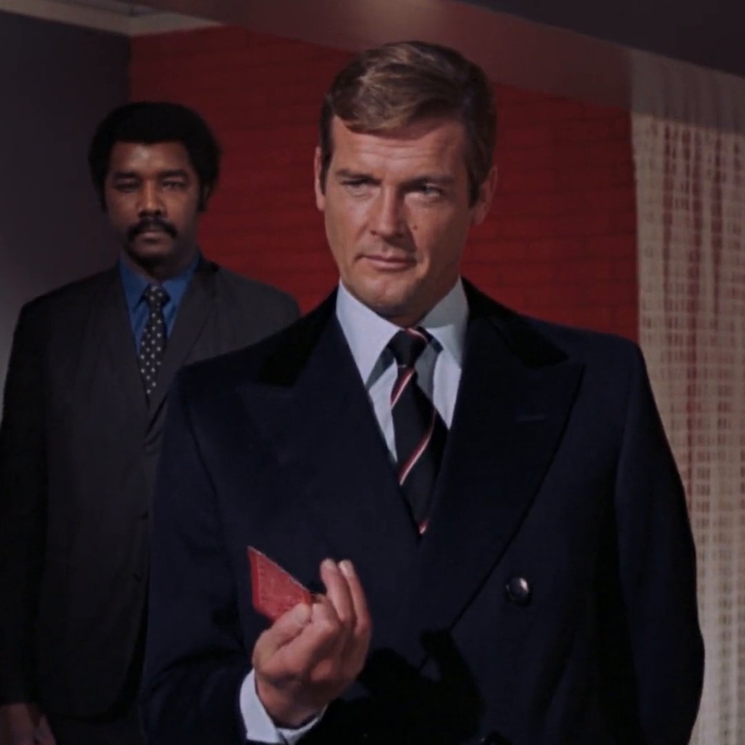 A still from a vintage film showing a man in a dark suit and striped tie holding a red card, with another man in a dark suit standing behind him.