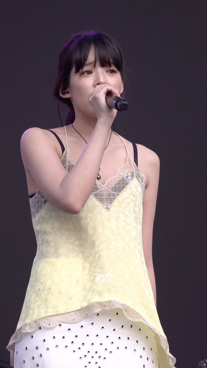 A young woman with dark hair and bangs performing on stage, wearing a pale yellow lace top and holding a microphone to sing.