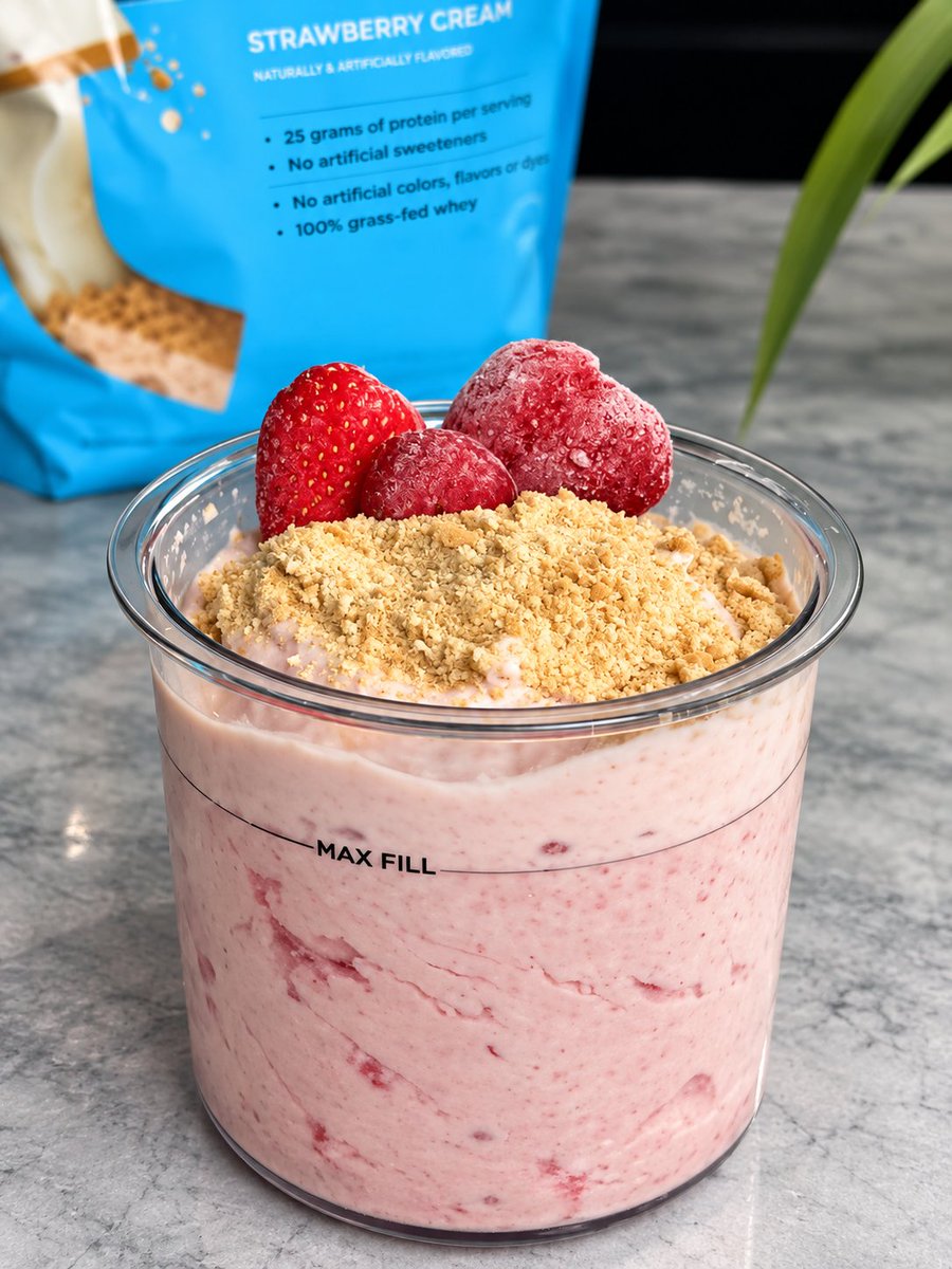 A clear plastic container marked 'MAX FILL' holds a pink strawberry cream mixture topped with graham cracker crumbs and three fresh strawberries, with a blue 'STRAWBERRY CREAM' protein powder bag (25 grams of protein per serving, 100% grass-fed whey) in the background.
