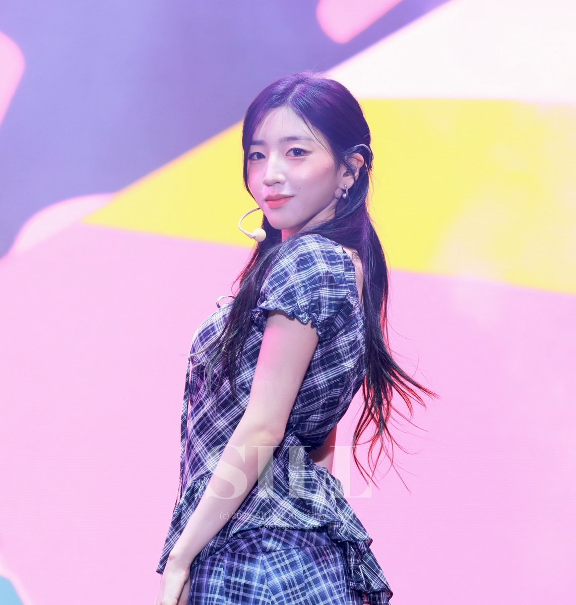 A young woman with long dark hair, wearing a blue plaid dress and a headset microphone, stands on a stage with a pink and yellow background; a 'JYP' watermark is visible.