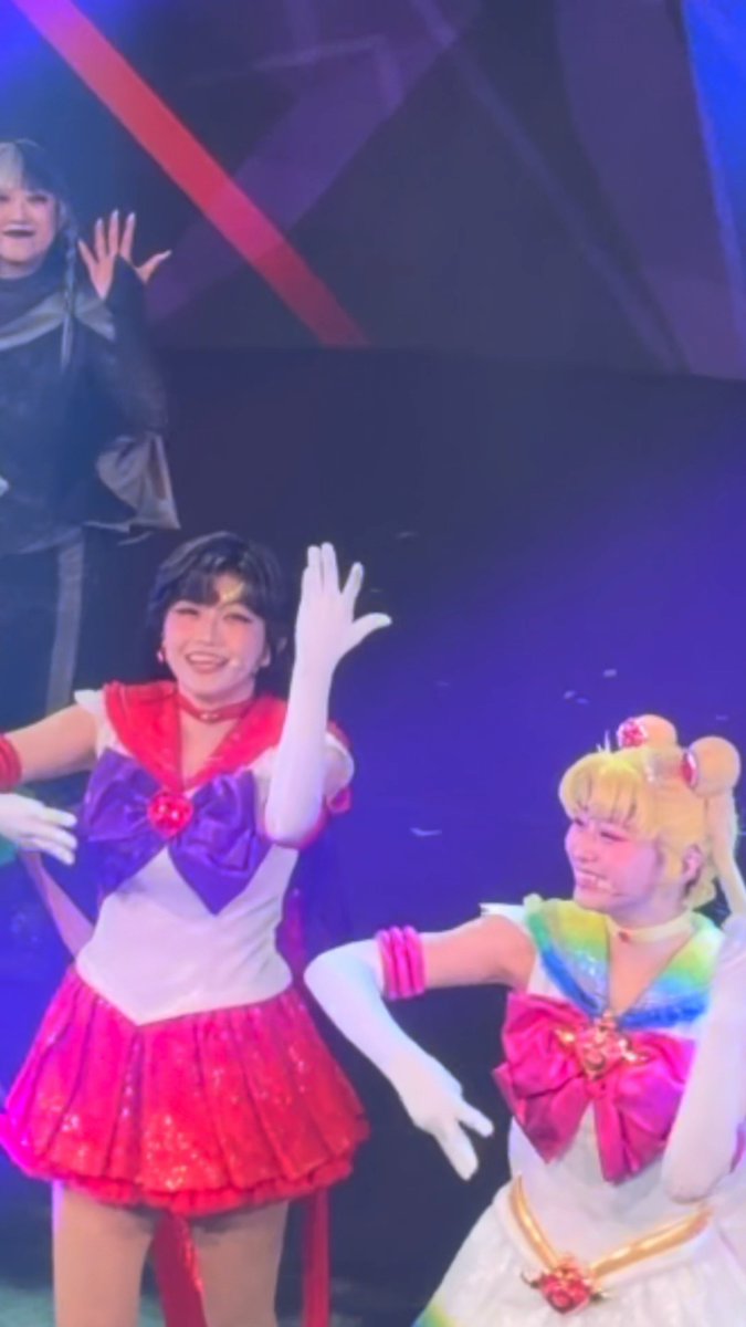 Two performers in Sailor Moon costumes on stage, with one in the classic red, white, and blue outfit waving and another in a rainbow-colored version of the costume.