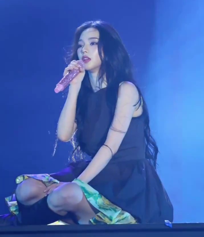 A female performer with long dark hair sings into a sparkly pink microphone while kneeling on stage under blue lighting, wearing a black sleeveless dress.