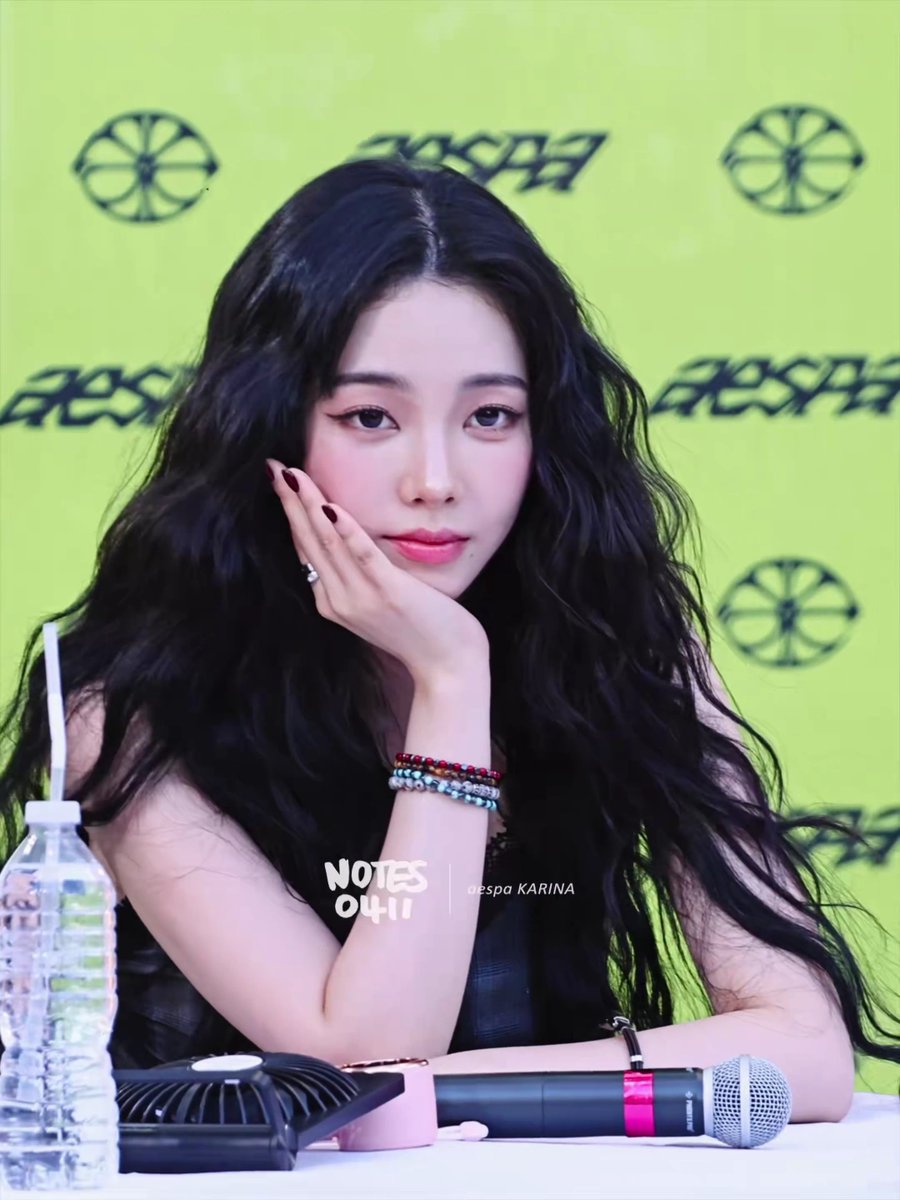 A young woman with long, wavy black hair rests her chin on her hand at what appears to be a fan event, with a neon green "aespa" branded backdrop behind her. The watermark reads "NOTES 04.11 | aespa KARINA," aligning with the "LA 카리나" caption indicating Karina of aespa during the group's LA appearance.