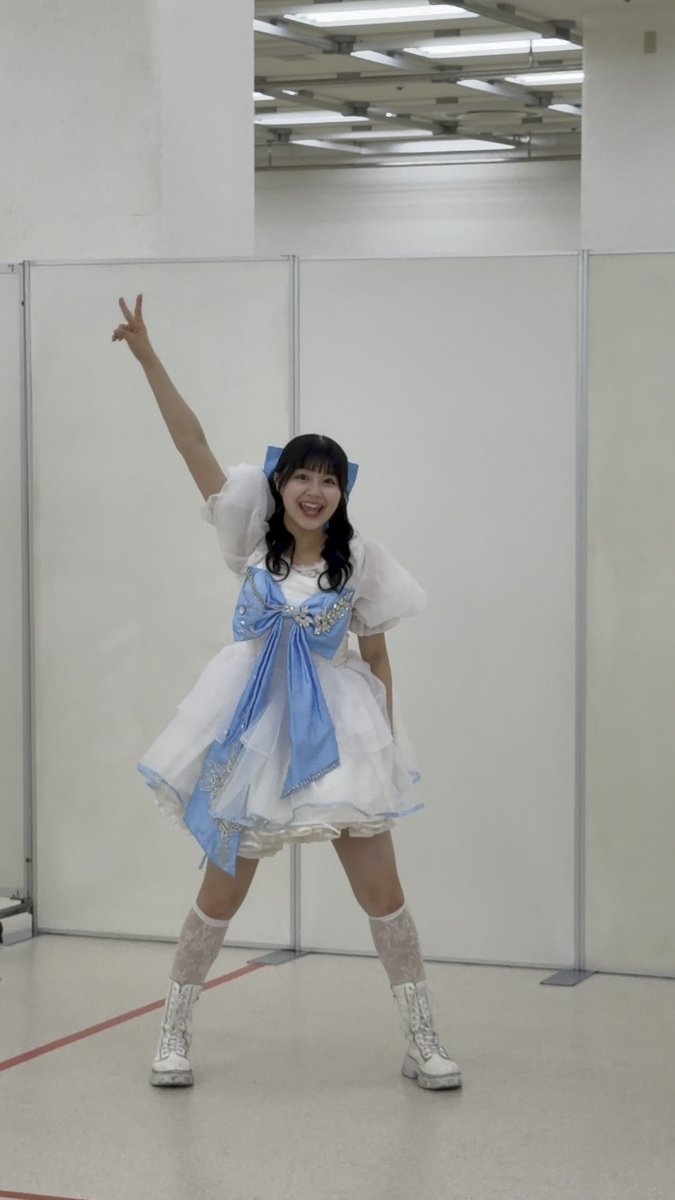 A woman in a frilly blue and white idol costume with knee-high lace socks and white boots poses with a peace gesture and a big smile.