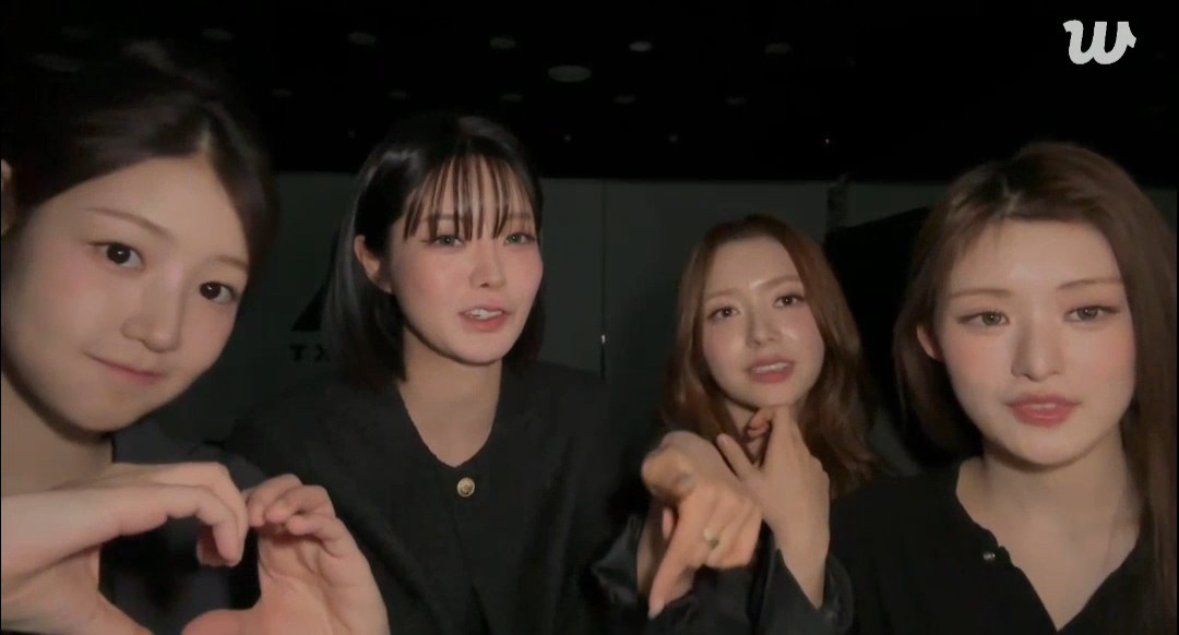 Four young women pose together indoors, with one making a small heart shape with her hands; a Weverse 'W' watermark appears in the top right corner.