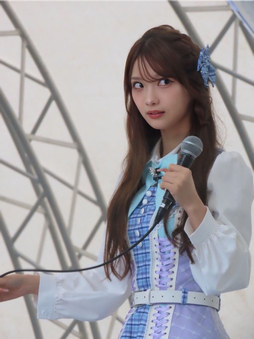 A young woman with long brown hair in braids holds a microphone while wearing a blue and white plaid outfit with lace-up details.