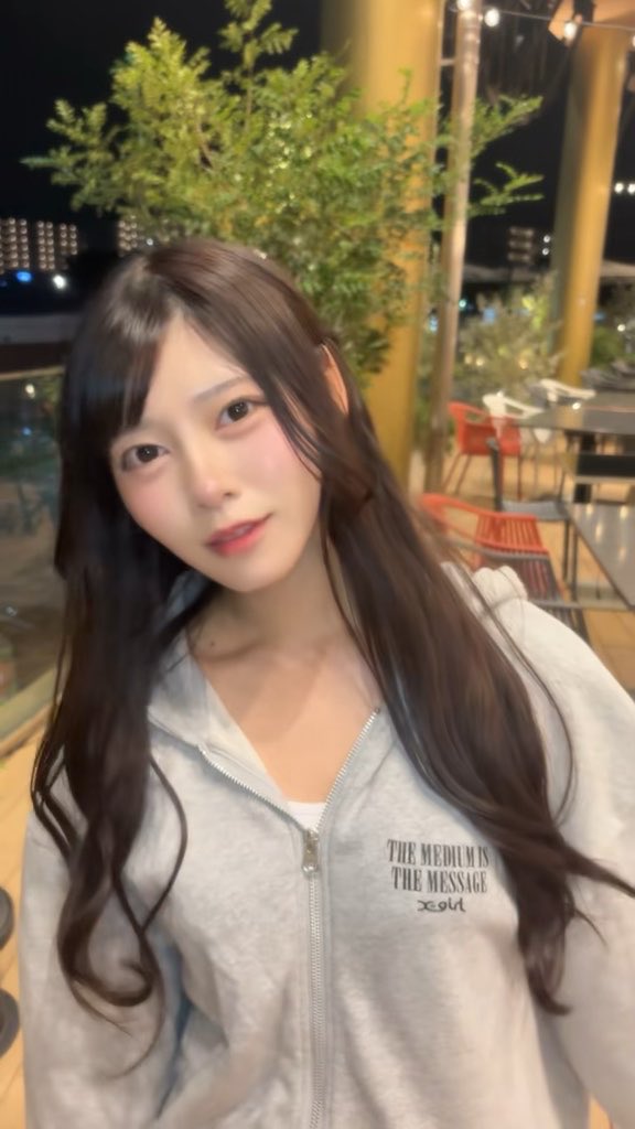 A young woman with long dark hair wearing a gray zip-up hoodie featuring the text "THE MEDIUM IS THE MESSAGE" poses for a selfie on an outdoor patio at night, likely promoting her updated TikTok account with the flirtatious caption "Do you like me?♡."