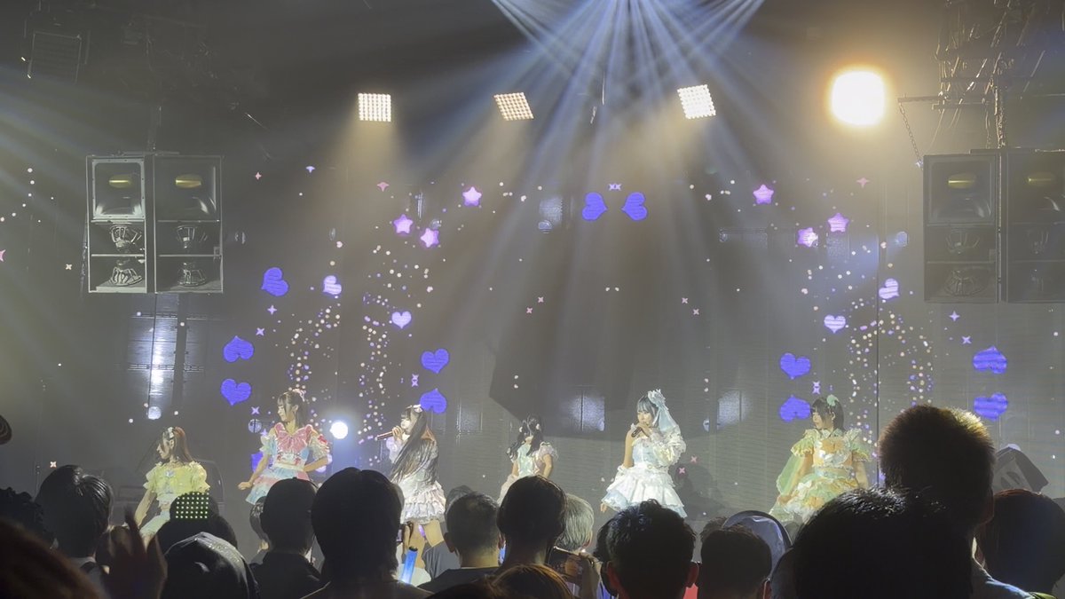 A group of performers in pastel dresses sing on a brightly lit concert stage decorated with star and heart projections, while an audience watches from below.
