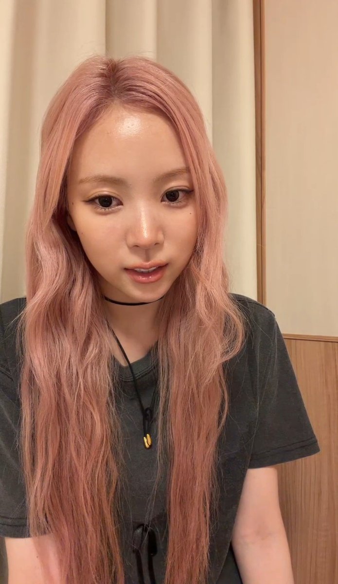 A young woman with long pink hair and a nose ring takes a selfie wearing a dark gray t-shirt and a pendant necklace.