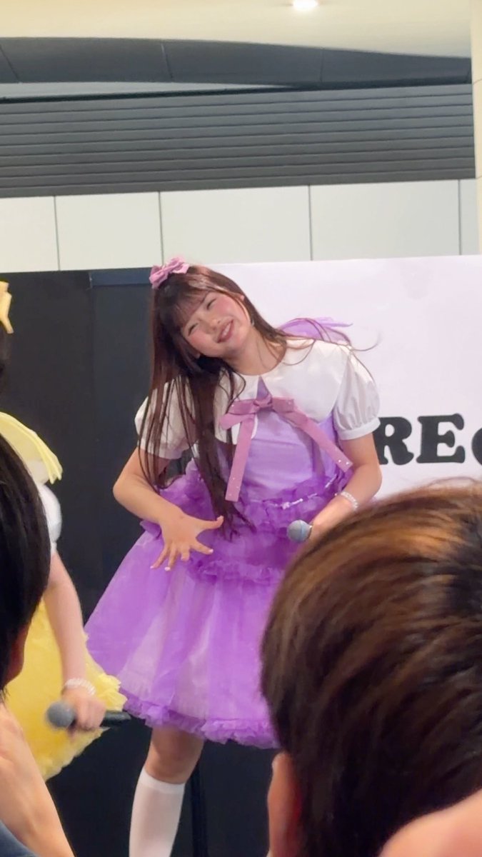 A young woman in a vibrant purple layered dress and white puff sleeves smiles brightly while performing on stage, with part of another performer visible to her left.