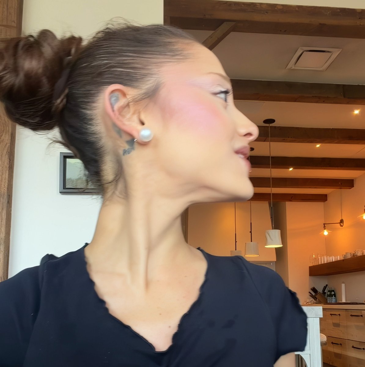 A profile view of a woman with dark hair in a high bun, wearing pearl earrings and a navy top, showing a small tattoo behind her ear in a kitchen with exposed wooden ceiling beams.