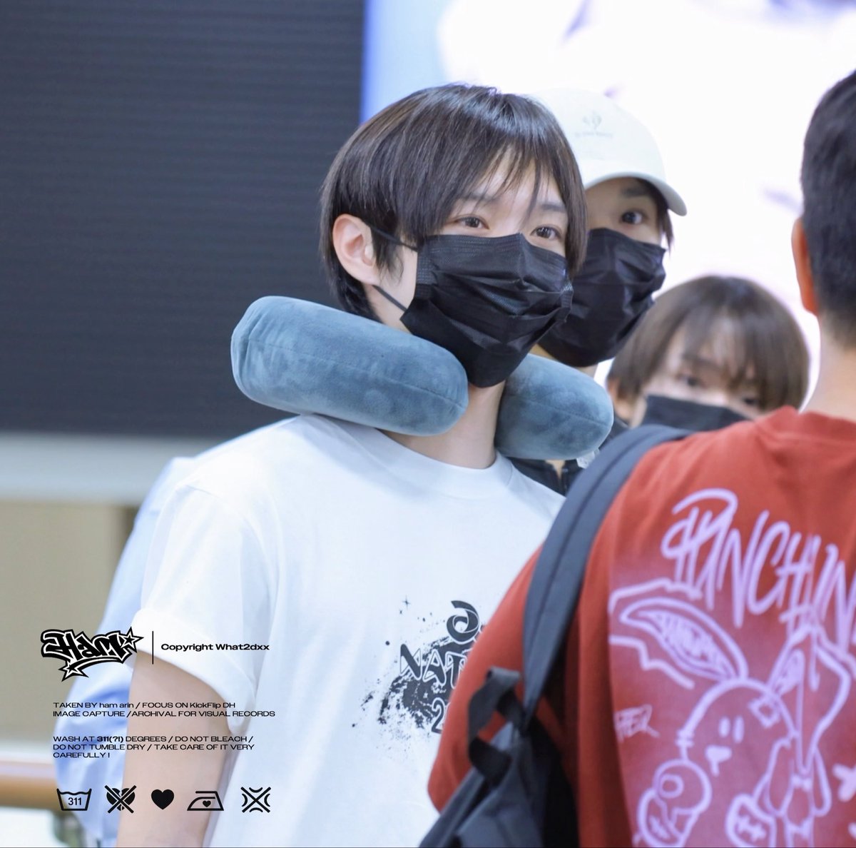 A young man wearing a black mask and a blue neck pillow is seen at what appears to be an airport, with a watermark reading 'Copyright What2dxx' and '2AM'.