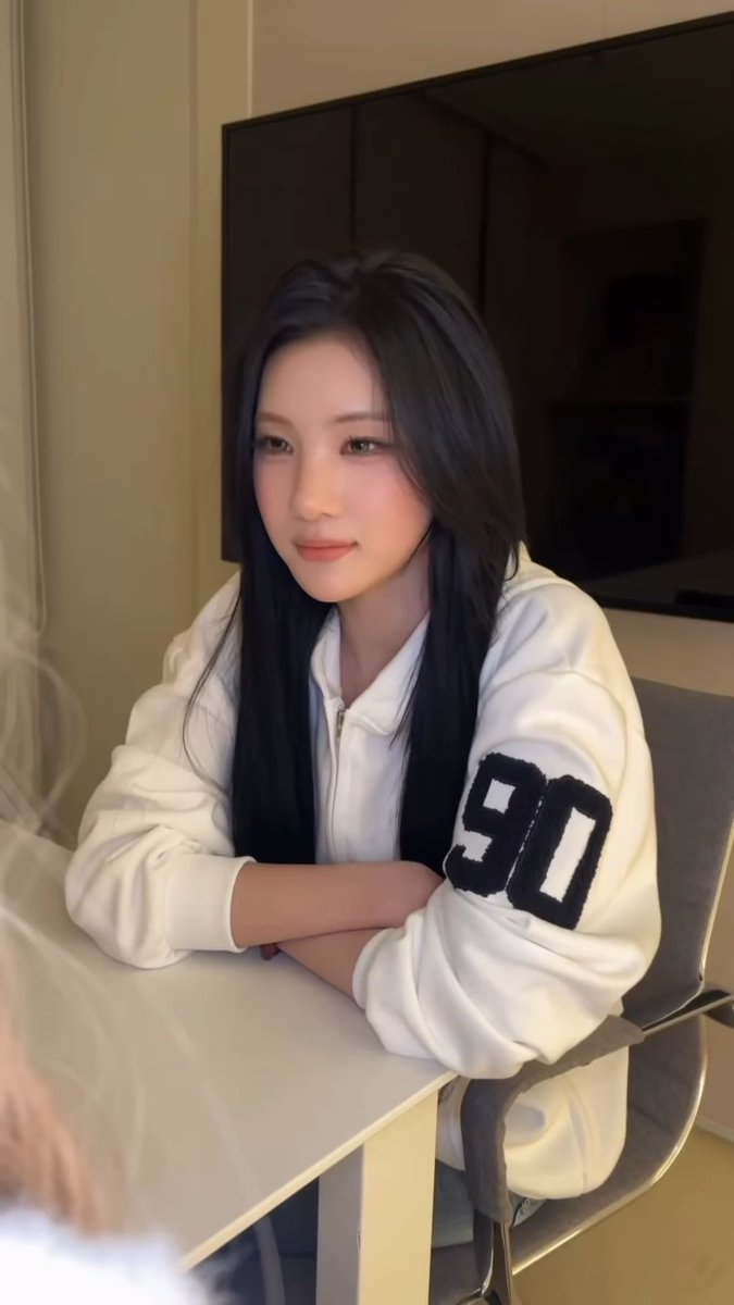 A young woman with long black hair sits at a white table wearing a white zip-up jacket with the number "90" on the sleeve, looking toward the camera in a softly lit room with a television in the background.