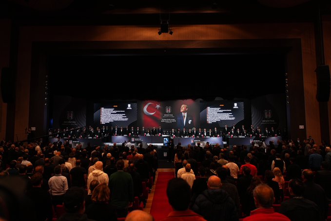 A crowded auditorium filled with attendees stands facing a stage during a Beşiktaş general assembly meeting, as displayed on the large screens showing the club's logo and "Olağan İdari ve Mali Genel Kurul" text, corresponding to the bjk.com.tr news report.