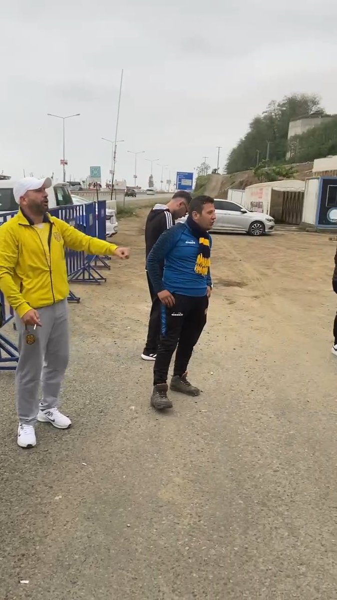 Outdoor scene showing a man in a yellow jacket and white cap pointing next to a man in a blue shirt at what appears to be a roadside location with barriers.